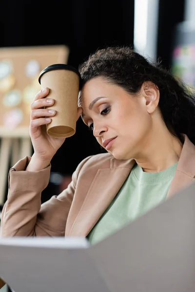 Втомилася афроамериканська бізнес-леді тримає паперову чашку біля голови, дивлячись на розмиту паперову теку — стокове фото