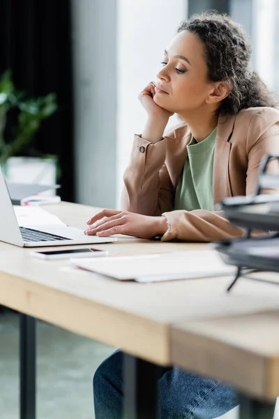 Вдумчивая американская бизнесвумен, сидящая на рабочем месте рядом с ноутбуком — стоковое фото