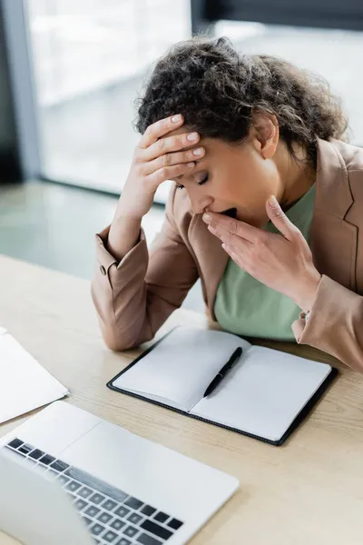 Müde afrikanisch-amerikanische Geschäftsfrau gähnt mit geschlossenen Augen neben leerem Notizbuch am Arbeitsplatz — Stockfoto