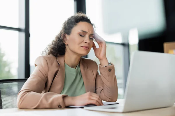 Agotada mujer de negocios afroamericana que sufre de dolor de cabeza cerca de la computadora portátil borrosa en el lugar de trabajo - foto de stock
