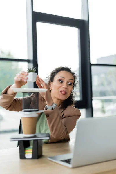 Mujer de negocios afroamericana sobresaliendo lengua mientras hace pirámide de papelería, taza de papel y maceta - foto de stock