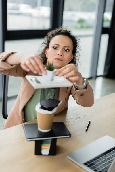 Mujer de negocios afroamericana con divertida mueca haciendo pirámide de maceta, papelería y taza de papel en el escritorio - foto de stock