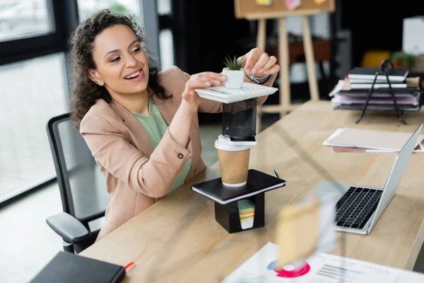 Mulher de negócios americana africana alegre fazendo pirâmide de artigos de papelaria, copo de papel e vaso de flores na mesa de trabalho — Fotografia de Stock