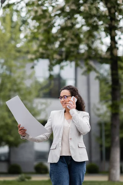 Positive african american businesswoman talking on smartphone while looking at papers on city street — Stock Photo