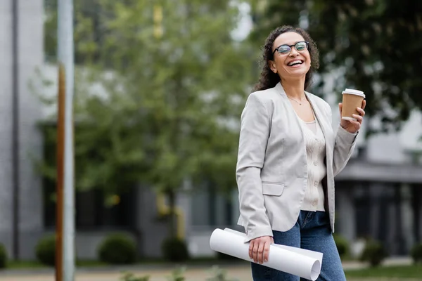 Веселая американская бизнесвумен держит кофе, чтобы пойти и свернуть бумаги на городской улице — стоковое фото