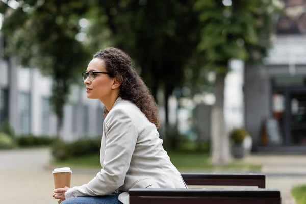 Side view of african american businesswoman in eyeglasses sitting with coffee to go outdoors — Stock Photo