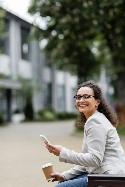 African american businesswoman with takeaway drink and mobile phone smiling at camera while sitting outdoors — Stock Photo