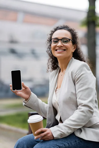 Happy african american businesswoman with paper cup showing smartphone with blank screen outdoors — Stock Photo