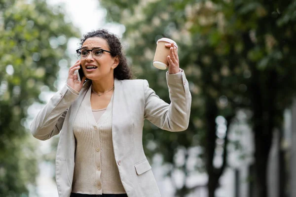 Preocupada mujer de negocios afroamericana con café para ir a hablar en el teléfono celular al aire libre - foto de stock