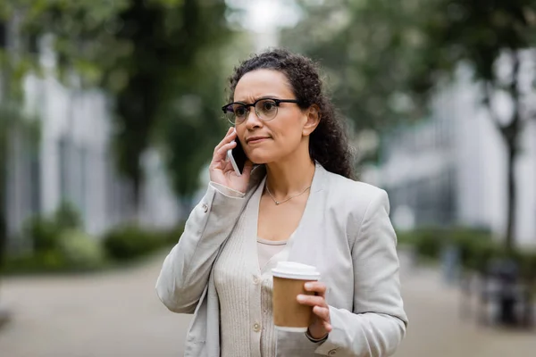 Nervous african american businesswoman with takeaway drink calling on mobile phone on city street — Stock Photo