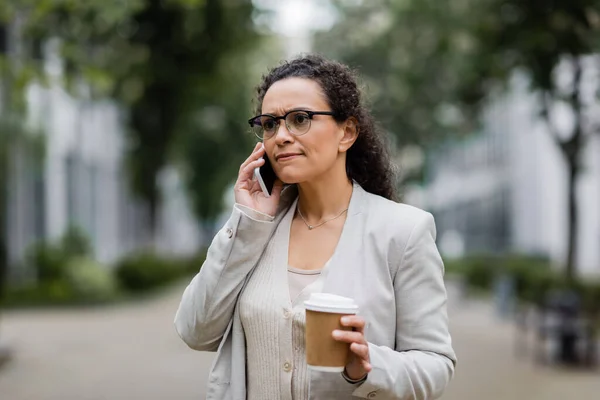 Nerviosa mujer de negocios afroamericana con bebida para llevar llamando por teléfono móvil en la calle de la ciudad - foto de stock