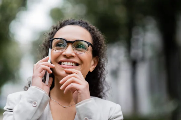 Mujer de negocios afroamericana feliz en gafas que hablan en el teléfono móvil al aire libre - foto de stock