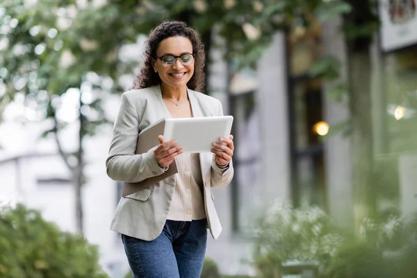 Donna d'affari afroamericana sorridente con documenti utilizzando tablet digitale su strada urbana — Foto stock