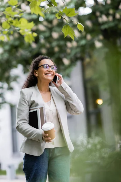 Happy african american businesswoman with documents and coffee to go talking on smartphone on city street — Stock Photo