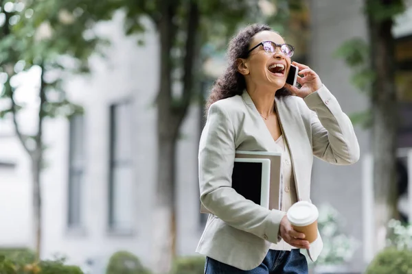 Laughing african american businesswoman with takeaway drink, digital tablet and documents talking on mobile phone outdoors — Stock Photo