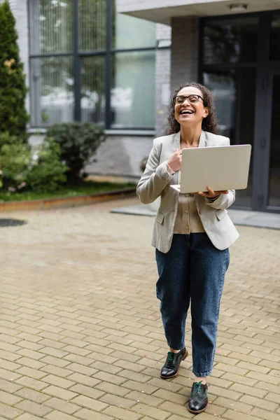 Full length view of cheerful african american businesswoman with laptop looking away near blurred building — Stock Photo