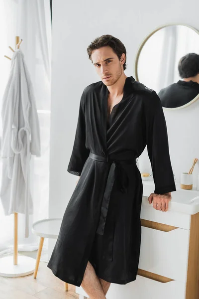 Young man in satin robe standing near sink in bathroom — Stock Photo