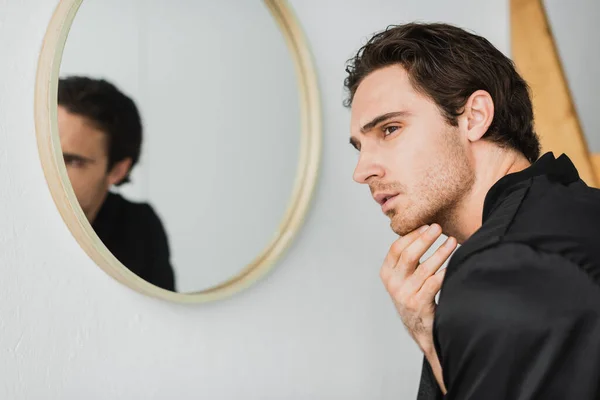 Jeune homme en robe de satin touchant le cou près du miroir dans la salle de bain — Photo de stock