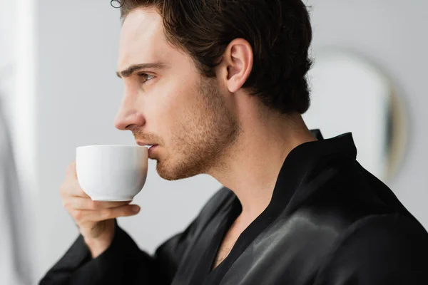 Side view of young man in robe holding coffee in bathroom — Stock Photo