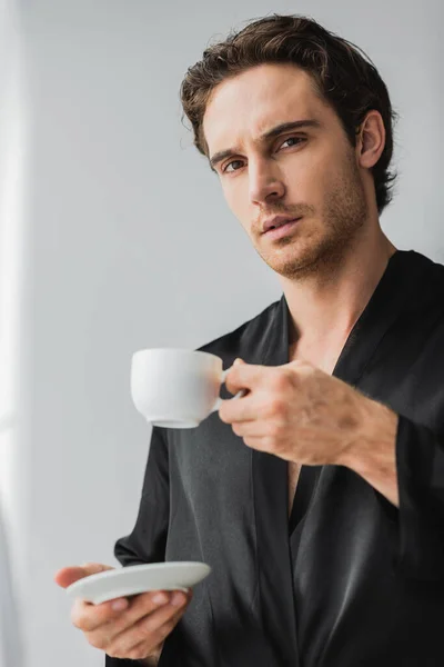 Young man in satin robe holding coffee cup with saucer and looking at camera at home — Stock Photo