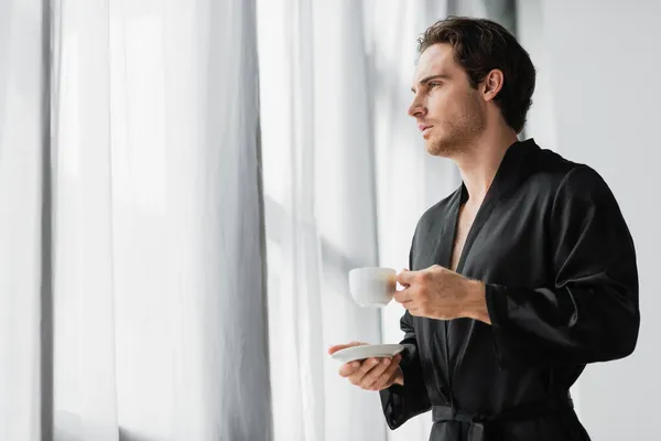 Joven en bata negra sosteniendo taza de café y platillo cerca de las cortinas en casa - foto de stock