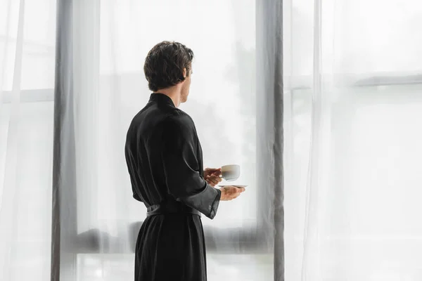 Homme en robe de soie noire tenant une tasse de café près des rideaux à la maison — Photo de stock