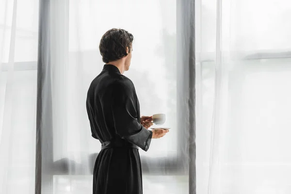 Homem em robe de seda preta segurando xícara de café perto de cortinas em casa — Fotografia de Stock