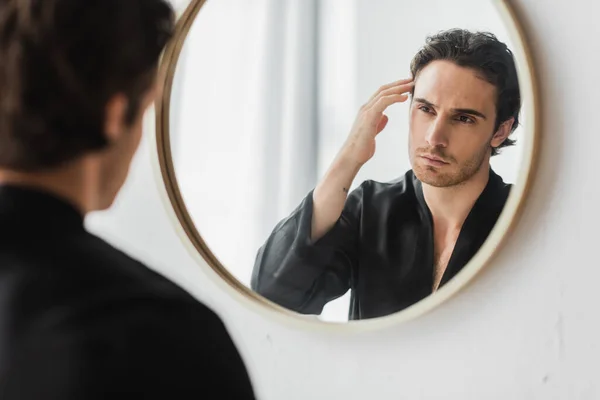 Hombre borroso en bata de seda tocando el cabello mientras mira el espejo en el baño - foto de stock