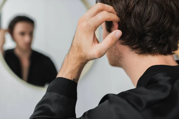 Jovem em robe de cetim tocando o cabelo perto do espelho borrado em casa — Fotografia de Stock