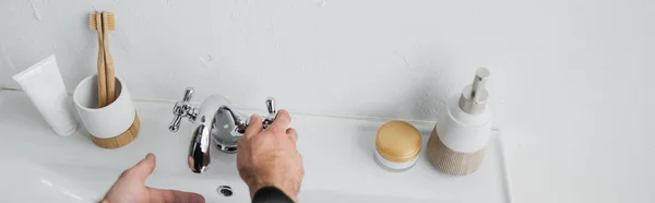 Ausgeschnittene Ansicht von Mann, der Wasser in der Nähe von Toilettenartikeln und Waschbecken im Badezimmer zapft, Banner — Stockfoto