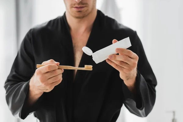 Cropped view of blurred man in robe holding toothbrush and toothpaste — Stock Photo