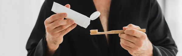 Vista recortada del hombre en bata sosteniendo pasta de dientes y cepillo de dientes en casa, pancarta - foto de stock