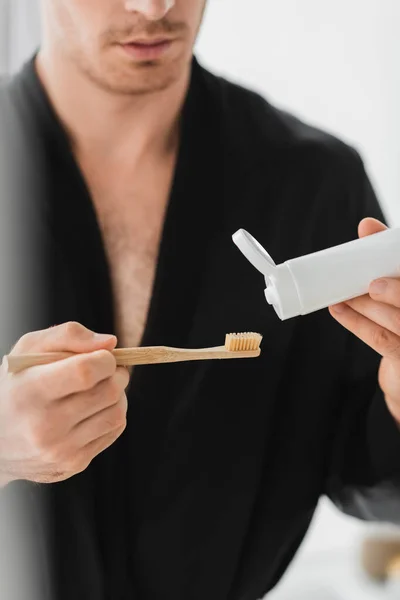 Vista recortada del hombre borroso en bata negra sosteniendo cepillo de dientes y pasta de dientes en el baño - foto de stock