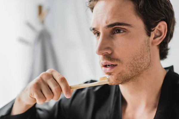 Young man in black robe holding wooden toothbrush with toothpaste in bathroom — Stock Photo