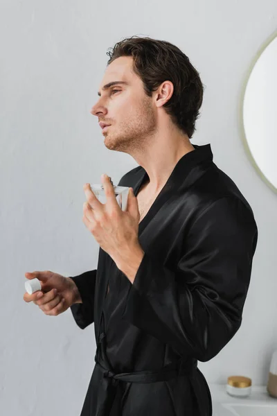 Hombre joven en bata de satén aplicando perfume en albornoz - foto de stock