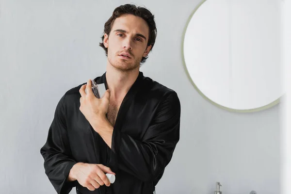 Young man in black robe applying perfume and looking at camera in bathroom — Stock Photo