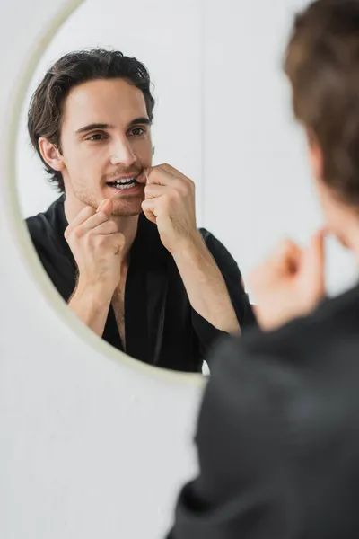 Uomo sfocato in accappatoio guardarsi allo specchio mentre pulisce i denti con filo interdentale — Foto stock