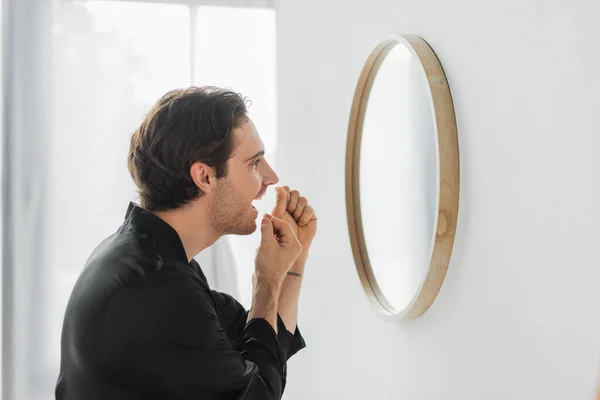Vista lateral del hombre en bata de satén con hilo dental cerca del espejo en el baño - foto de stock