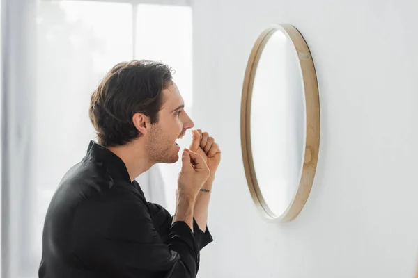 Vista laterale dell'uomo in accappatoio di raso utilizzando filo interdentale vicino specchio in bagno — Foto stock