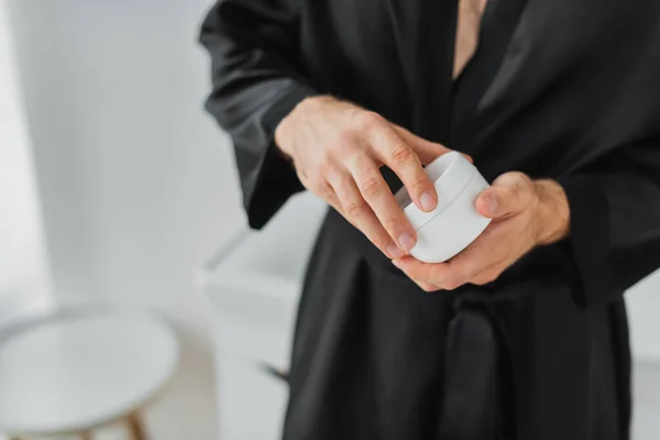 Vue recadrée de l'homme en robe de satin noir tenant pot avec crème cosmétique dans la salle de bain — Photo de stock