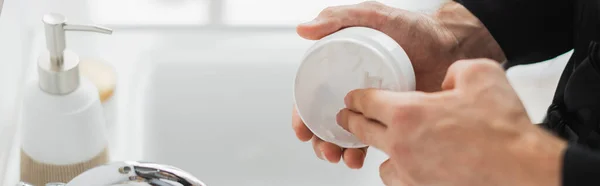 Ausgeschnittene Ansicht des Mannes, der kosmetische Creme aus dem Glas in der Nähe des Wasserhahns im Badezimmer nimmt, Banner — Stockfoto