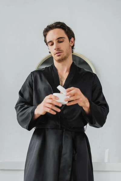 Jeune homme en robe de satin tenant pot flou avec crème cosmétique dans la salle de bain — Photo de stock