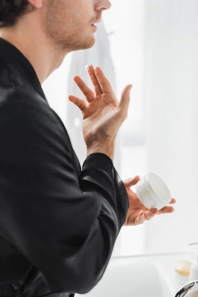 Cropped view of young man in silk robe holding cosmetic cream in bathroom — Stock Photo