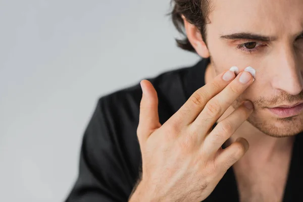 Vue recadrée du jeune homme appliquant de la crème sur le visage isolé sur le gris — Photo de stock