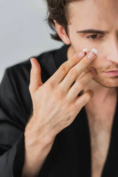 Vista recortada del joven en bata negra aplicando crema cosmética en la cara aislada en gris - foto de stock