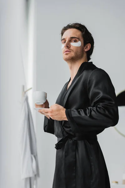 Low angle view of man in silk robe with eye patches holding cup of coffee in bathroom — Stock Photo