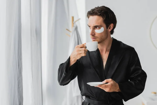 Hombre joven en bata negra y parches para los ojos sosteniendo la taza de café en el baño - foto de stock