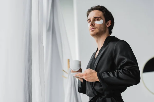 Vista basso angolo di uomo in accappatoio di raso e patch per gli occhi che tengono il caffè vicino tende in bagno — Foto stock