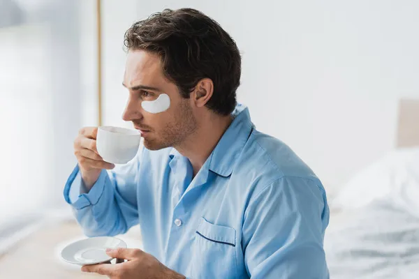 Side view of young man in pajama and eye patch drinking coffee in bedroom — Stock Photo