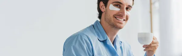 Hombre positivo en pijama y parches para los ojos sosteniendo una taza de café en casa, pancarta - foto de stock
